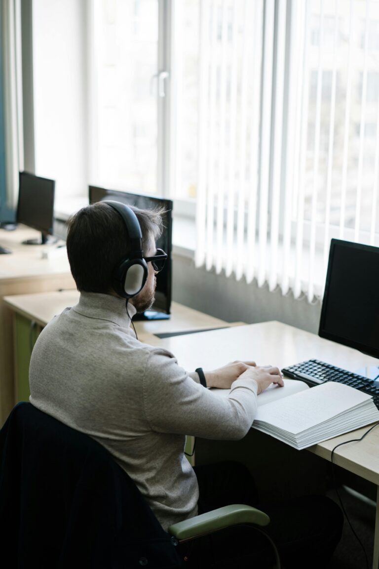 A blind man using headphones reads a Braille book at an office desk with computers.