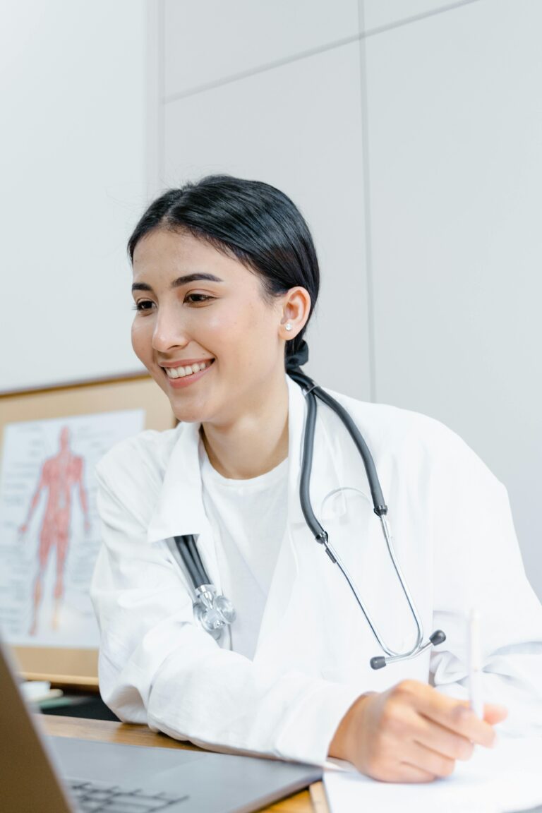 Confident female doctor in a lab coat smiling during an online consultation using a laptop.